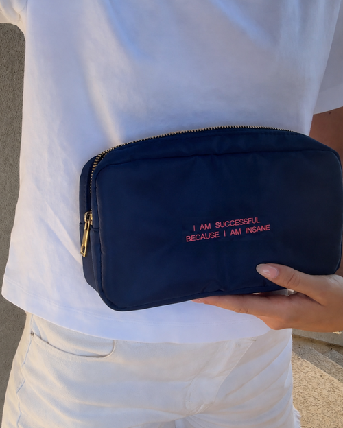 A navy blue nylon cosmetic pouch with gold zipper and pink embroidered text reading 'I AM SUCCESSFUL BECAUSE I AM INSANE' held by a person wearing a white t-shirt and white pants.